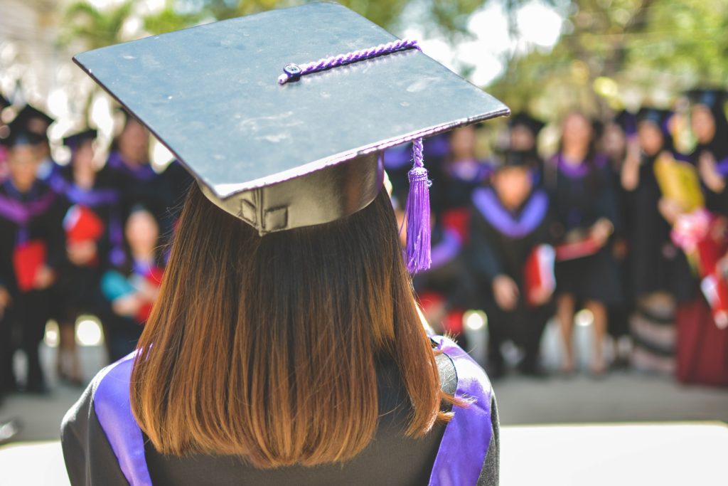 Woman with brown hair with back to camera wearing black graduation cap and gown and facing other graduates